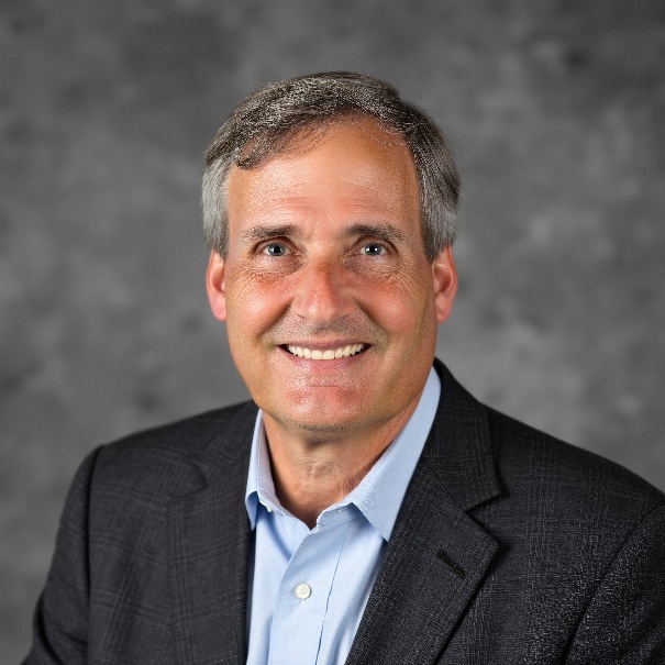 Headshot of Warren B. Powell — silver-grey hair, navy blazer, light blue shirt, smiling, photographed against a grey background