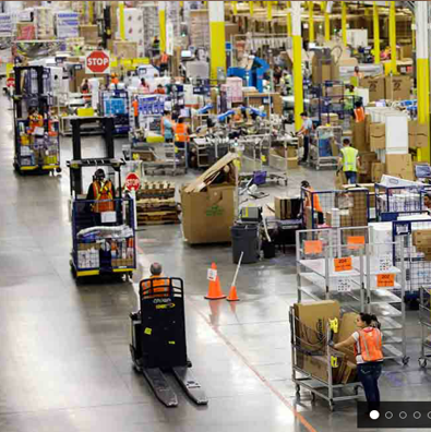 A wide-angle photo of the interior of a large Amazon fulfillment center, with workers in orange vests pushing wheeled carts down long aisles of merchandise