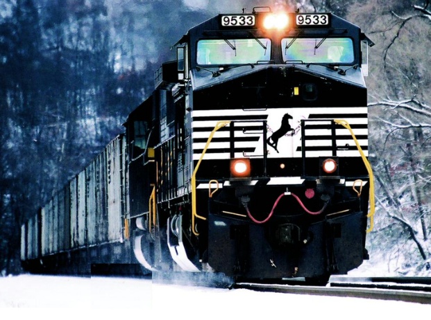 Front view of a Norfolk Southern locomotive number 9533 emerging from a snowy forest, with the railroad's white-horse logo on the front