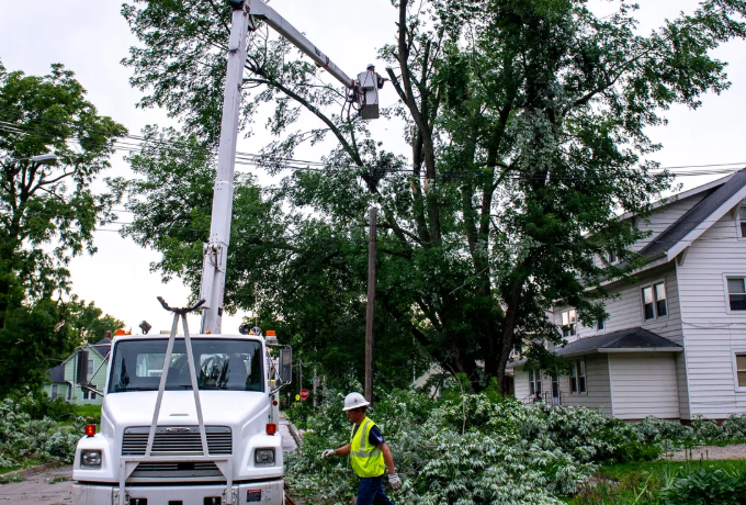 A utility truck with a cherry-picker boom positioned next to a tree with damaged limbs, with a utility worker on the ground assessing the damage after a storm